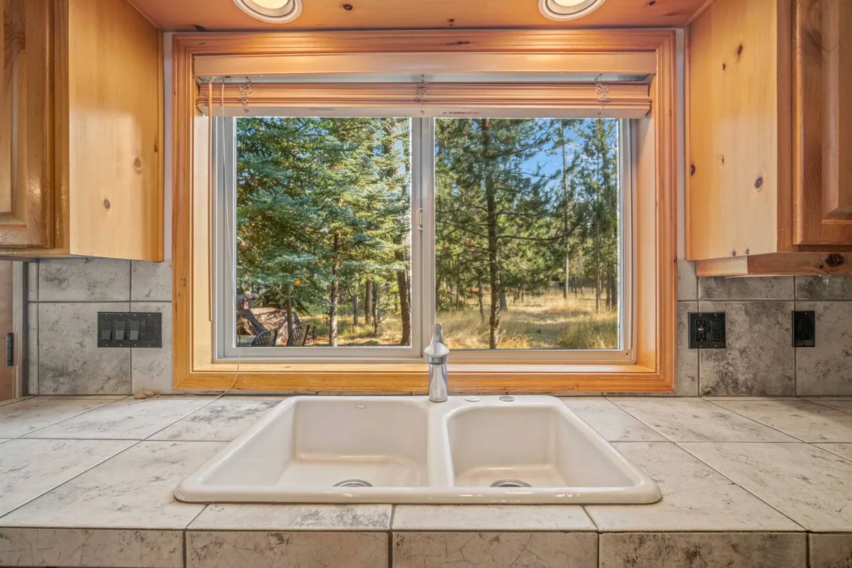 A cozy kitchen scene featuring a sink in the foreground, with a large window behind it letting in natural light and offering views of the outdoors. The activities suggested include enjoying nature while cooking or entertaining, and the mood is warm and inviting.
