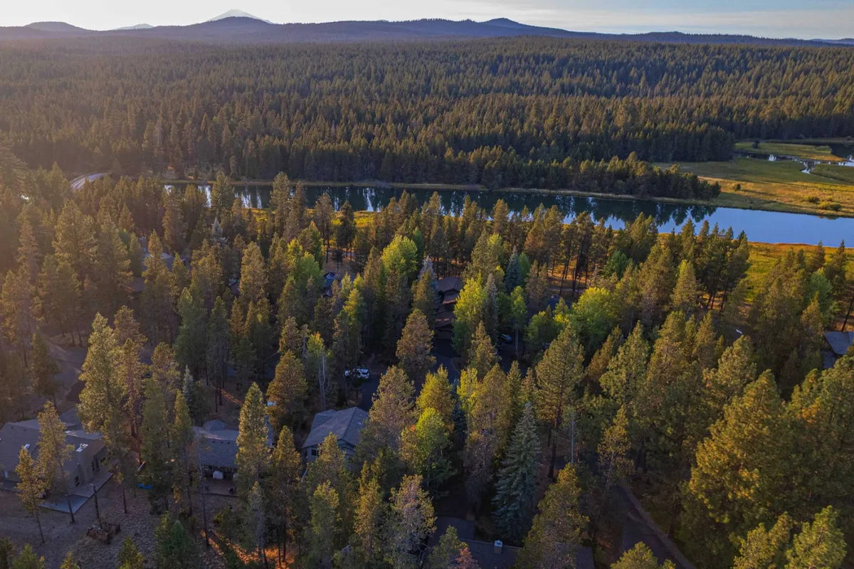 A scenic exterior view where dense forest surrounds a gentle river, distant mountains, and homes tucked among the trees, emphasizing the natural beauty and serene setting of the Sunriver area.