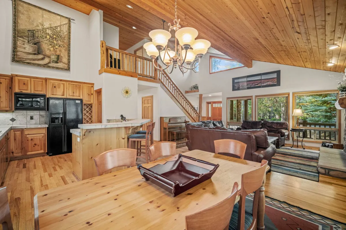 A bright, welcoming dining area with a wooden ceiling, large windows that overlook the outdoors, a central dining table, and a chandelier. The image conveys a scene of family gathering, relaxation by the fireplace, and morning coffee with nature views.
