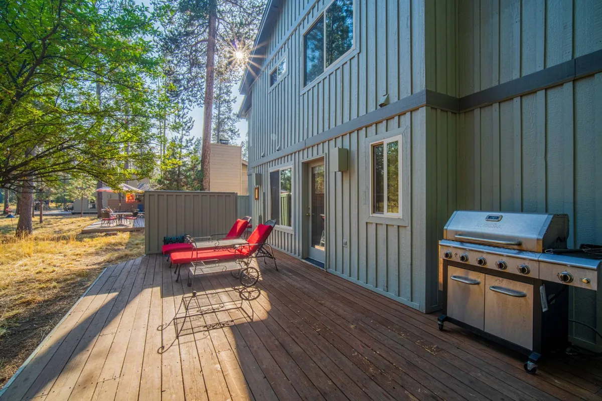 This image shows a spacious wooden deck surrounded by tall pine trees, featuring two vibrant red lounge chairs and a sleek stainless steel BBQ grill. The atmosphere is casual and welcoming, with ample space for socializing or relaxing outdoors. The deck is bathed in natural sunlight, reinforcing the connection to the serene natural setting.