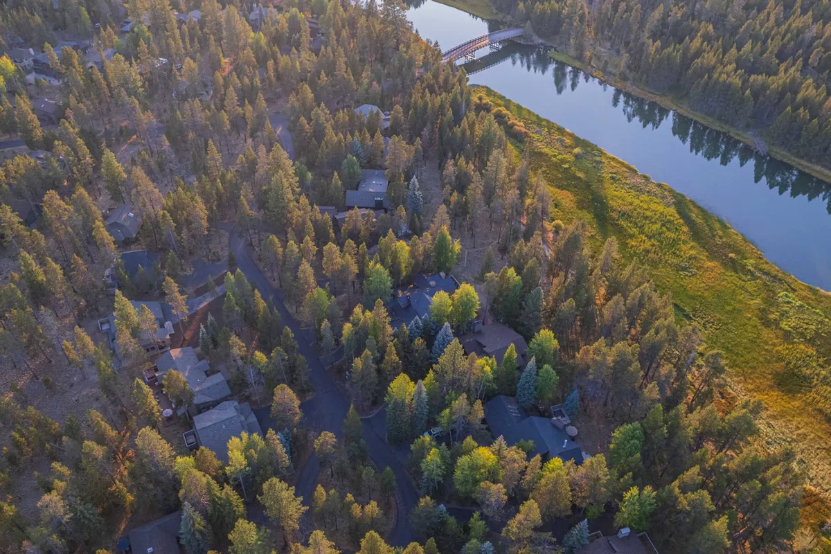 A serene river flows beneath a bridge surrounded by lush forest and a cluster of homes, illustrating easy access from the property to natural waterways and trails. The setting sun casts warm reflections on the water, highlighting the area's calm and inviting atmosphere.