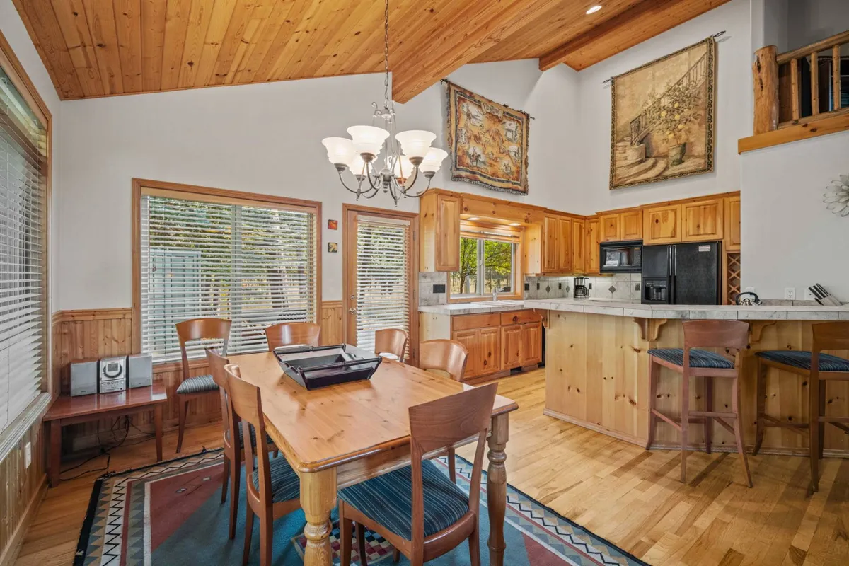 A bright, inviting dining area featuring a large table set for a meal, a stylish chandelier overhead, a kitchen island in the background, and large windows with tree views. The space feels tailored for sharing stories and celebrating milestones.