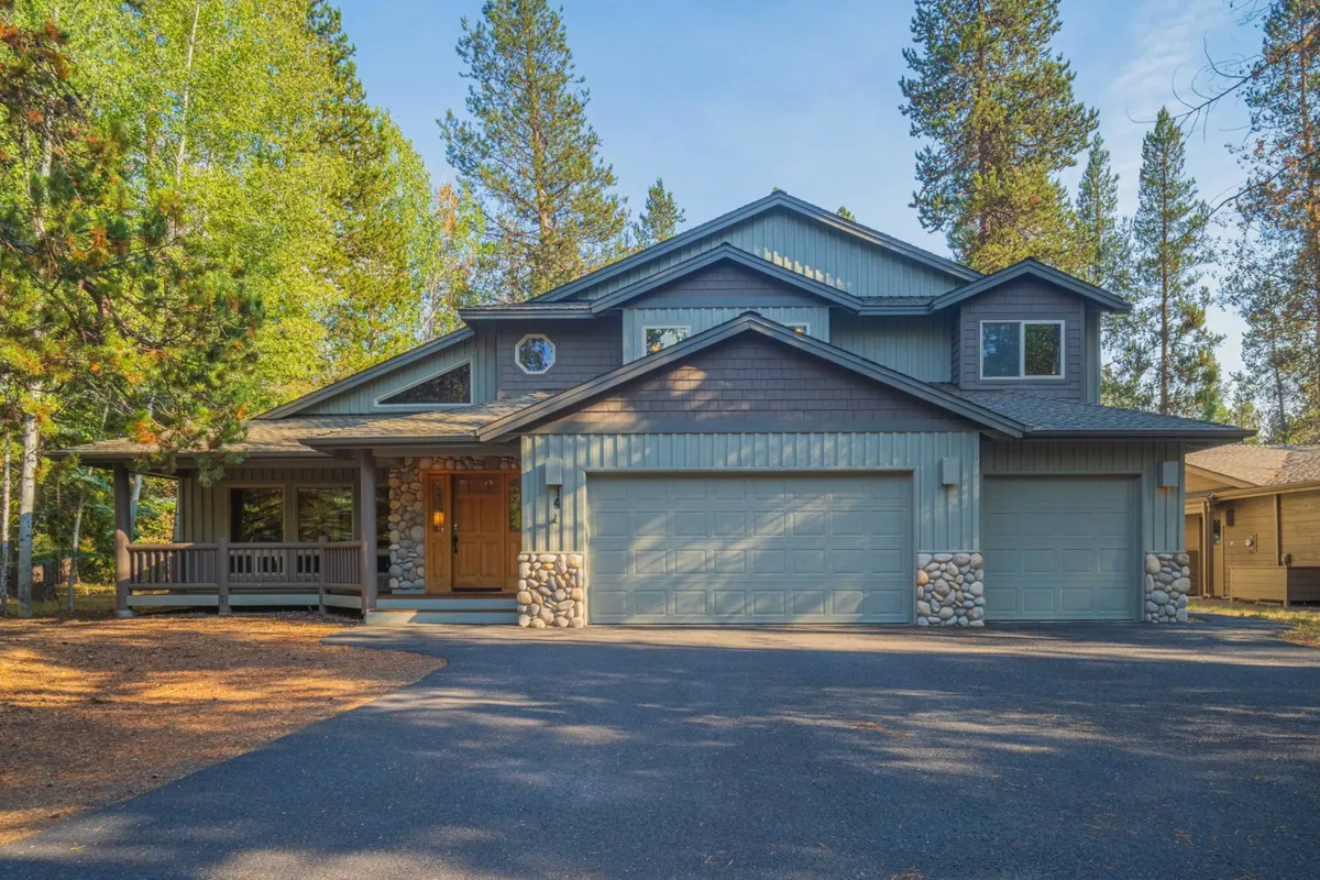 This exterior shot features the front porch of the home, highlighted by stone accents and large garage doors. The driveway is visible, flanked by tall windows that hint at the spacious interior. The porch is inviting, surrounded by mature trees, perfect for family gatherings and setting off on biking adventures.