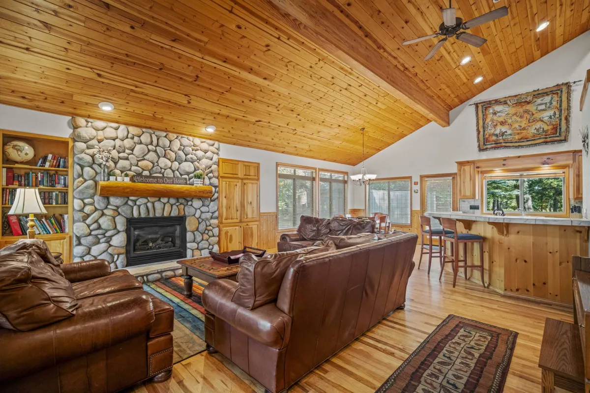 The image showcases a spacious, inviting living area featuring a stunning stone fireplace as the centerpiece, high wooden ceilings, plush leather sofas, a built-in bookshelf, and a modern kitchen bar in the background. The space exudes warmth and comfort, offering multiple spots for relaxation, conversation, and cozy gatherings.