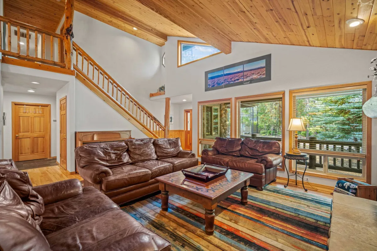 A cozy living room scene with leather sofas, a colorful area rug, large windows for natural light, and a wooden staircase. The space is arranged for socializing, relaxing, or sharing stories after a day out.