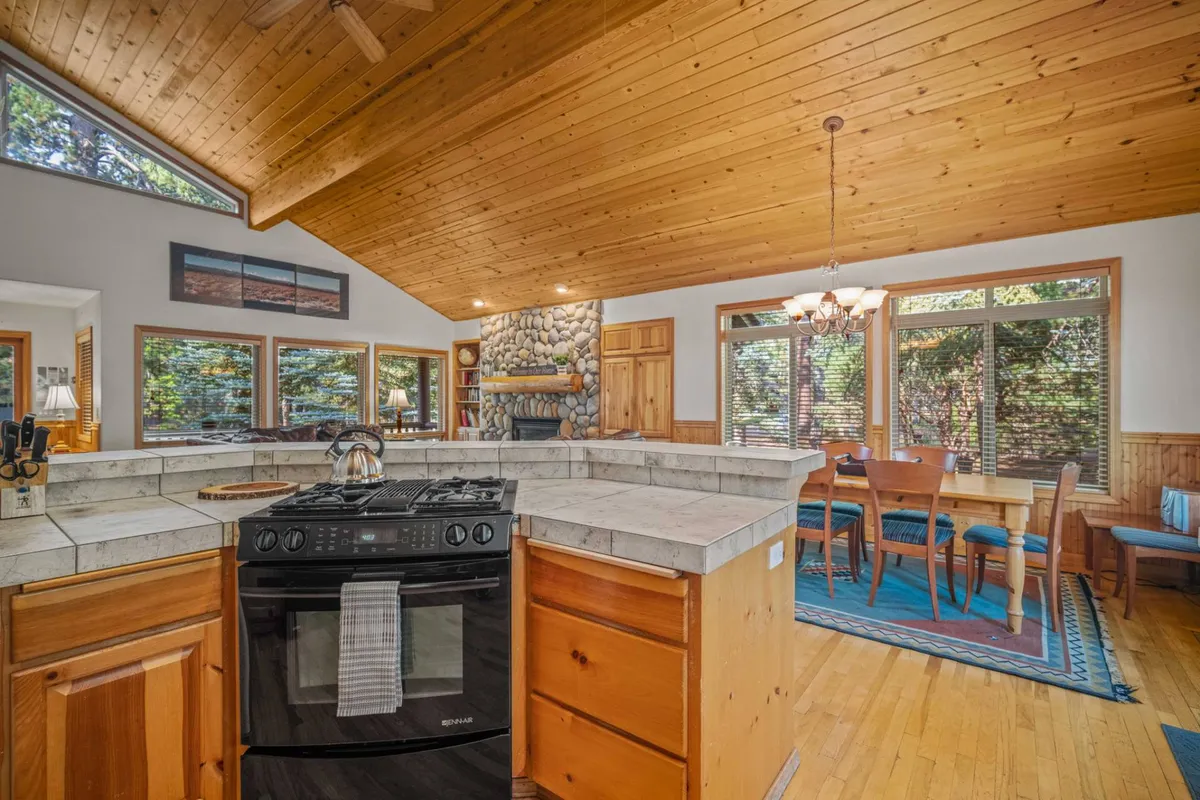 This image shows a warm, open-concept living space that connects the kitchen, dining, and living room. The focal points include a kitchen island, a prominent stone fireplace, large windows letting in natural light, and a cozy dining area. Leather sofas and a soft area rug add comfort, while the open layout encourages togetherness.
