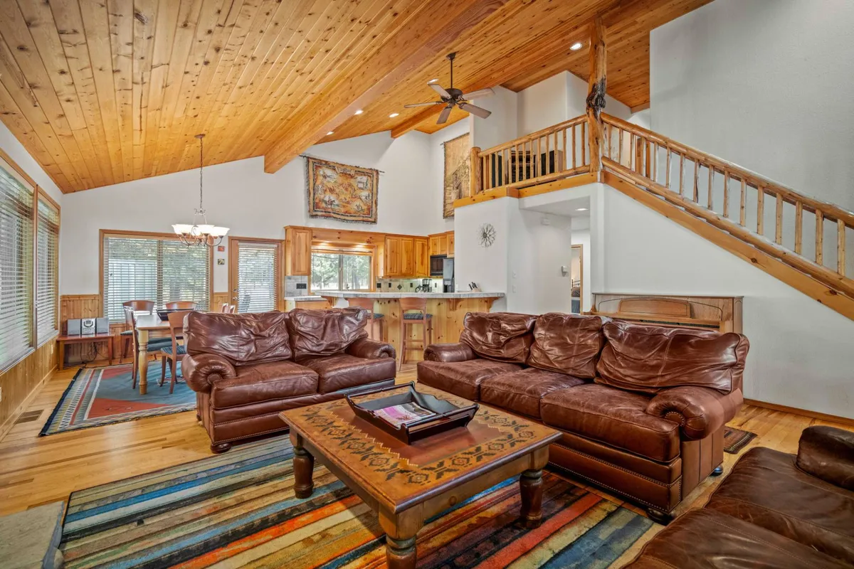 A wide view of the living room featuring plush leather sofas, a central coffee table, wooden ceiling accents, and a visible dining area and kitchen beyond. The atmosphere is warm and welcoming, with ample space for family gatherings and relaxation.