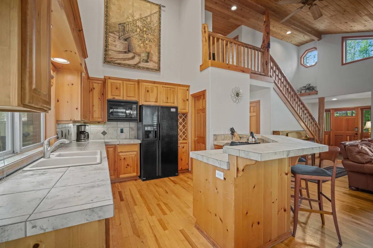 Focused on the kitchen, this image presents a kitchen island, black refrigerator, rich wooden cabinetry, and a large window that floods the space with light. The staircase and open sightlines suggest easy flow to surrounding living areas, while the kitchen feels upgraded and ready for family feasts.